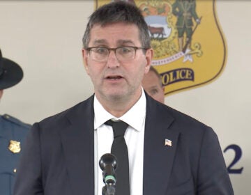 Delaware Governor Matt Meyer addresses members of the media, speaking into a microphone at a podium, with a Delaware state trooper behind him.