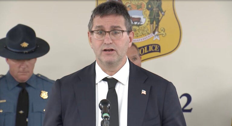 Delaware Governor Matt Meyer addresses members of the media, speaking into a microphone at a podium, with a Delaware state trooper behind him.