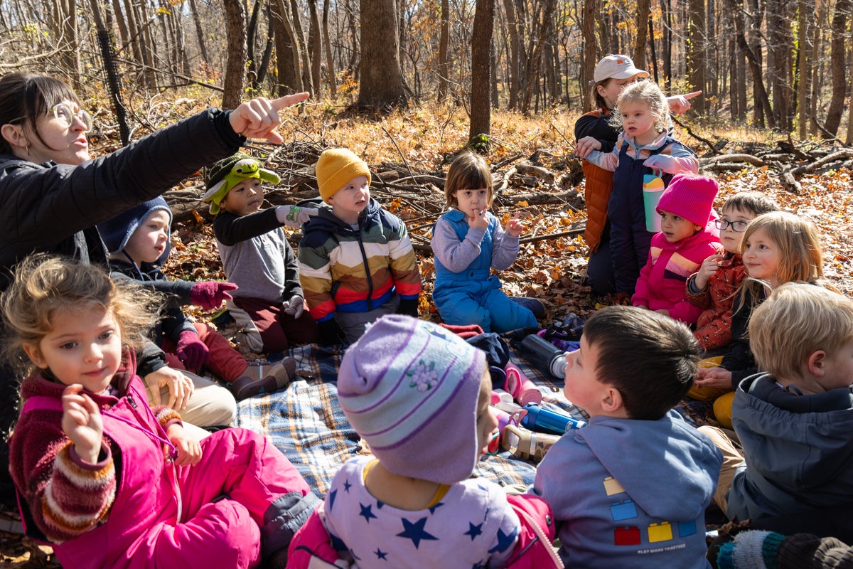 Children huddle in a circle outside while their teacher points at something to look out of frame