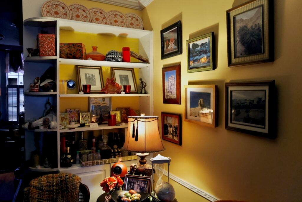 A lamp illuminates a dark corner of a home, with a full bookshelf, and many framed photographs on the wall.