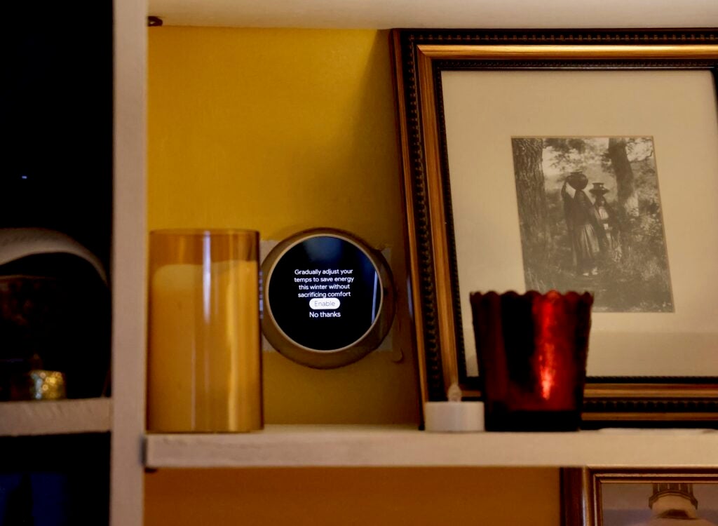 A smart thermostat is on the wall above a shelf with a candle and framed photo on it.