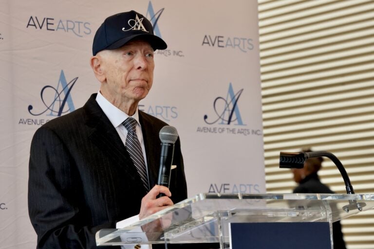 Carl Dranoff holding a microphone as he stands at a podium with an Avenue of the Arts backdrop