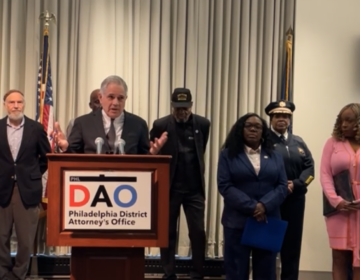 Larry Krasner speaking with many people behind him in front of a podium with a Philadelphia District Attorney's Office sign