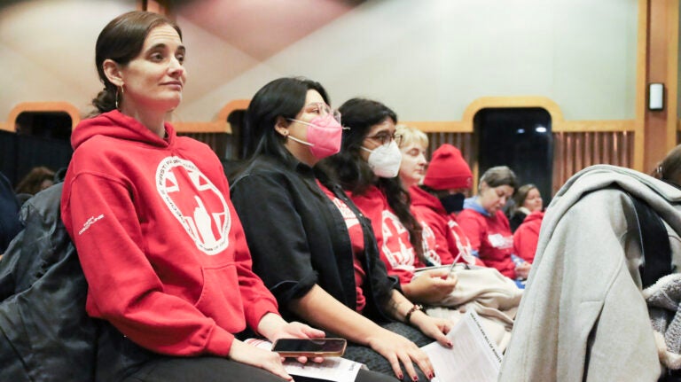 Health care workers and advocates sit in a forum, most wearing the same red sweater