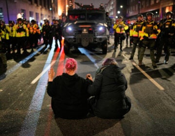 Two people sit in the street holding hands in front of Minnesota State Patrol during a protest
