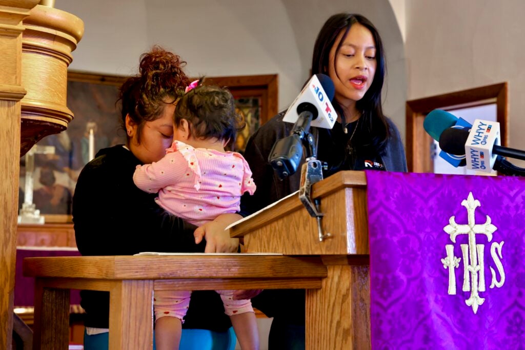 Ashley Batz reads a statement at a podium while Andrea holds her young daughter