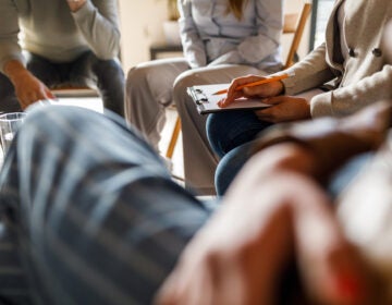 A therapist holding a clipboard for notes on her lap during a group therapy session.