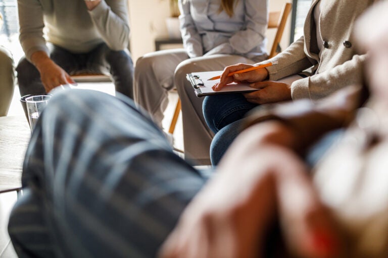A therapist holding a clipboard for notes on her lap during a group therapy session.