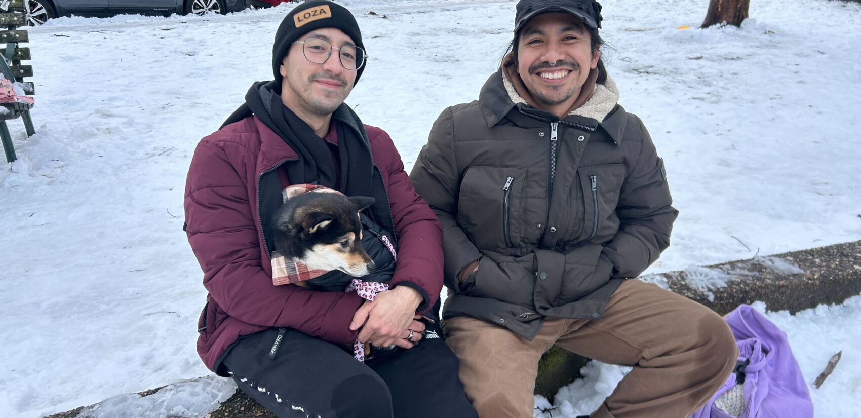 Two people and their dog pose for a picture while sitting on a log in the park after the snowstorm