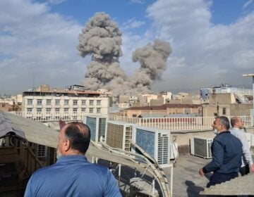 People watch as smoke rises on the skyline after an explosion in Tehran, Iran