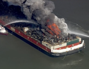 a large barge is billowing smoke from a fire, while 2 small ships spray it with water to put the fire out.