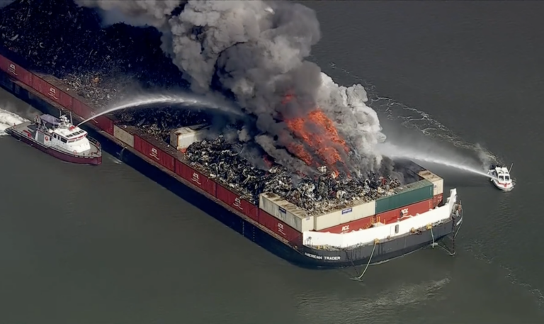 a large barge is billowing smoke from a fire, while 2 small ships spray it with water to put the fire out.