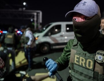 A masked Immigration and Customs Enforcement agent holds a baton