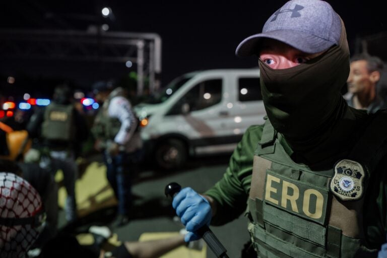 A masked Immigration and Customs Enforcement agent holds a baton