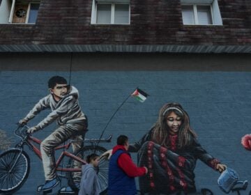 People are walking on the street in front of a large mural depicting a child on a bike with a Palenstinian flag