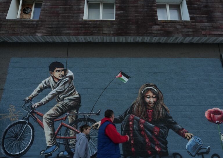 People are walking on the street in front of a large mural depicting a child on a bike with a Palenstinian flag