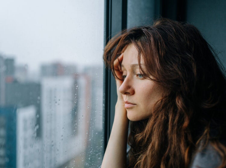 Young woman with brown hair stares through a rain-streaked window in a city apartment, pensive and melancholic, reflecting on loneliness and mental health during autumn A woman with brown hair looks sadly out of a window