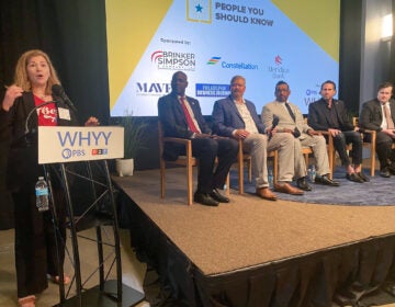 sports-economy-panel-j-mellman-2026-05-21 Maiken Scott is standing at a podium next to a stage which features five panelists sitting in a row.