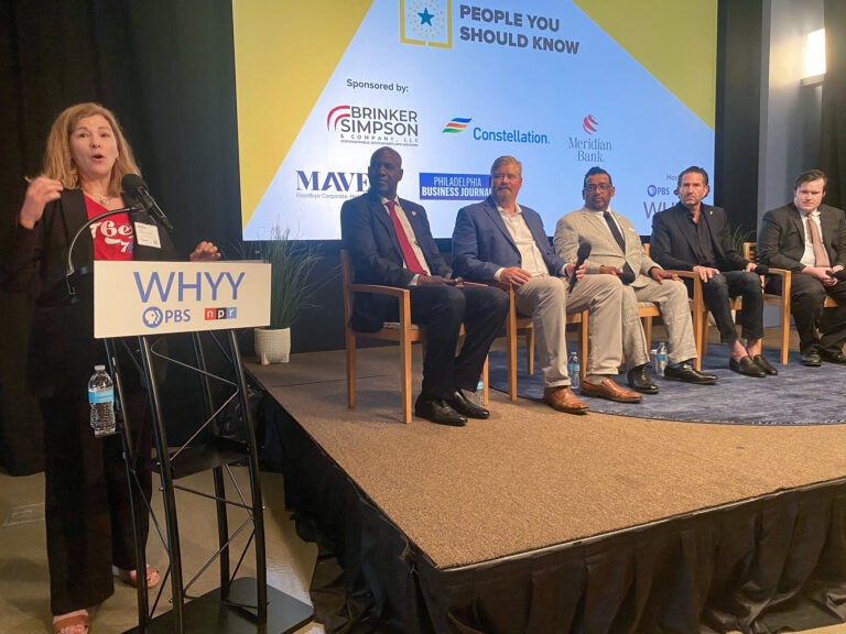 Maiken Scott is standing at a podium next to a stage which features five panelists sitting in a row.