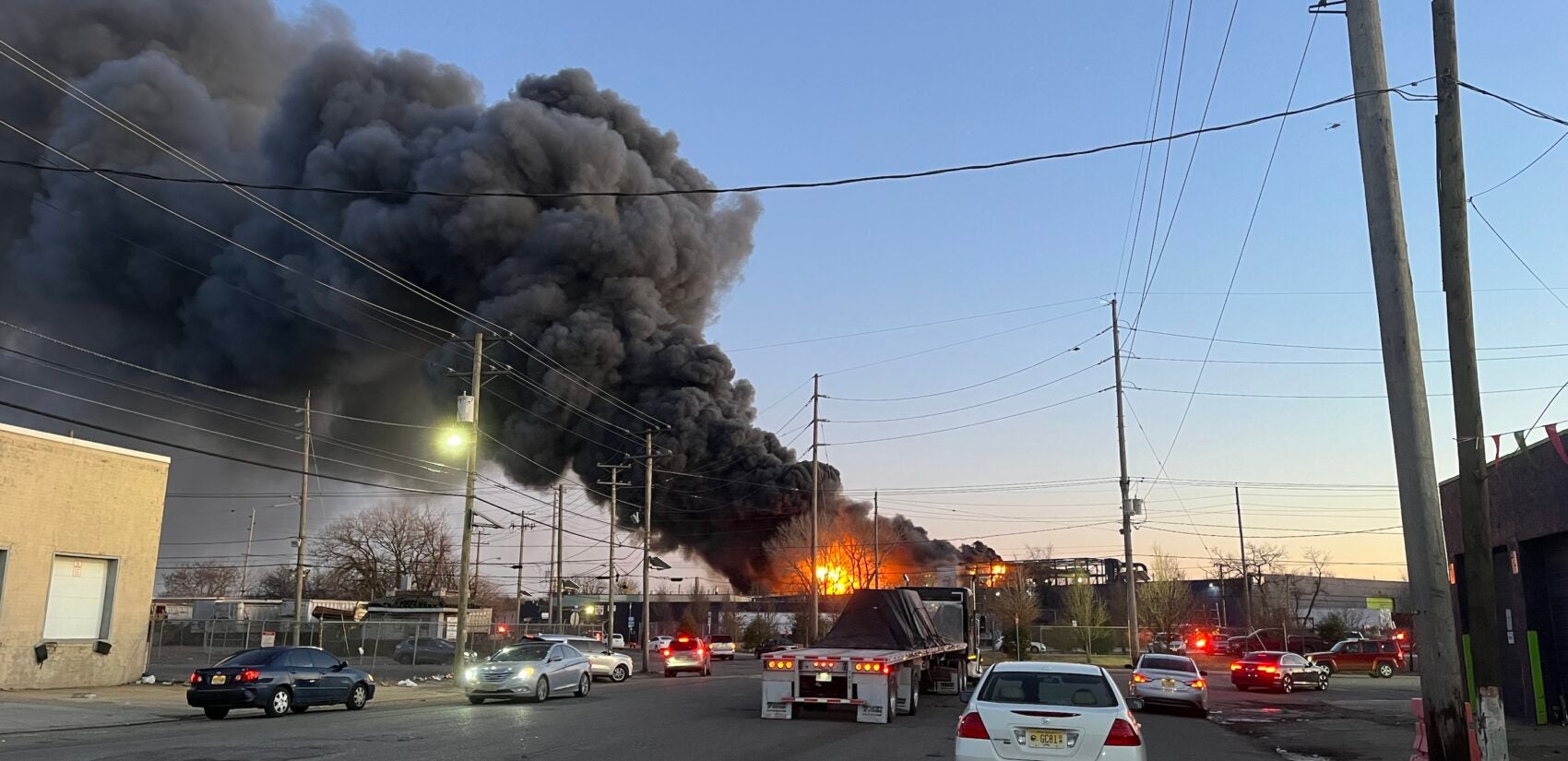 A fire rages in the distance as cars drive down the road.