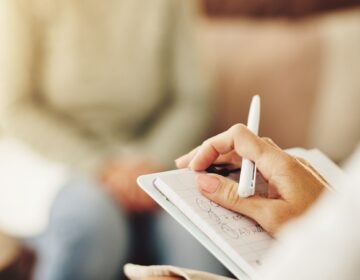 The hands of someone holding a pen and notepad while talking to someone else are seen.