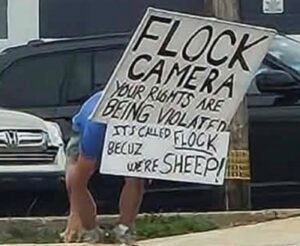 Joshua Pyle holds a sign reading FLOCK CAMERA YOUR RIGHTS ARE BEING VIOLATED IT'S CALLED FLOCK BECUZ WE'RE SHEEP!