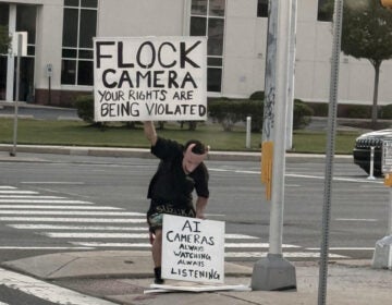 flock-protest-signs-millcreek-delaware-2026-cropped Joshua Pyle, wearing a mask inspired by the character Lock from the movie Nightmare Before Christmas, holds two signs, one reading FLOCK CAMERA YOUR RIGHTS ARE BEING VIOLATED, and the other reading AI CAMERAS ALWAYS WATCHING ALWAYS LISTENING