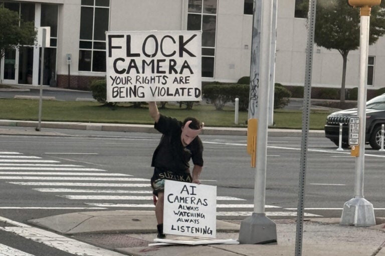 Joshua Pyle, wearing a mask inspired by the character Lock from the movie Nightmare Before Christmas, holds two signs, one reading FLOCK CAMERA YOUR RIGHTS ARE BEING VIOLATED, and the other reading AI CAMERAS ALWAYS WATCHING ALWAYS LISTENING