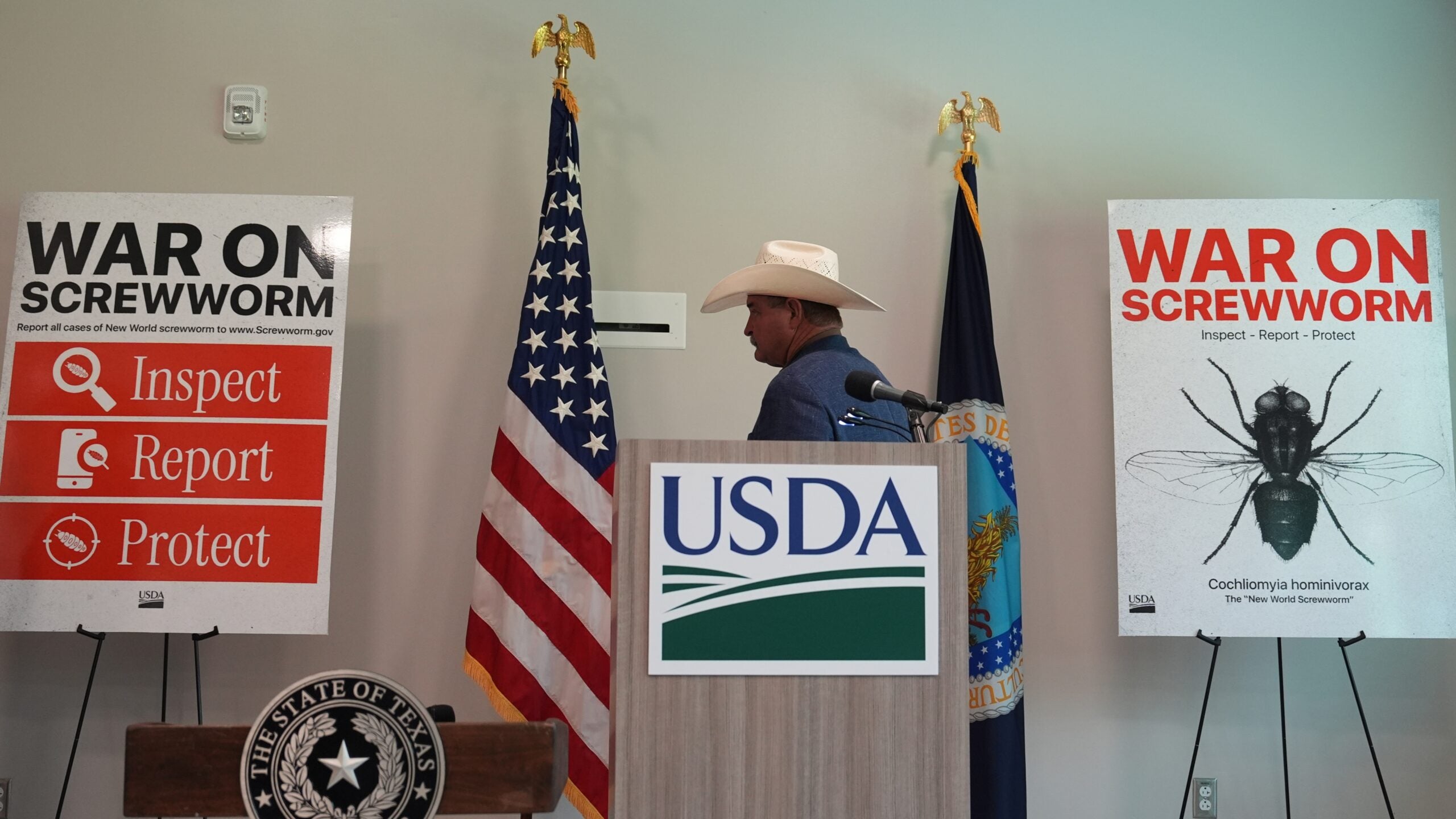 a man stands between two signs reading WAR ON SCREWWORM INSPECT REPORT PROTECT