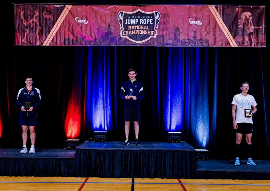Zach Peckman is standing at the top and in the center of a three-tiered podium at the Jump Rope championship. He is smiling as he holds up his gold medal. The second and third place winners are on either side of him on shorter levels of the podium.
