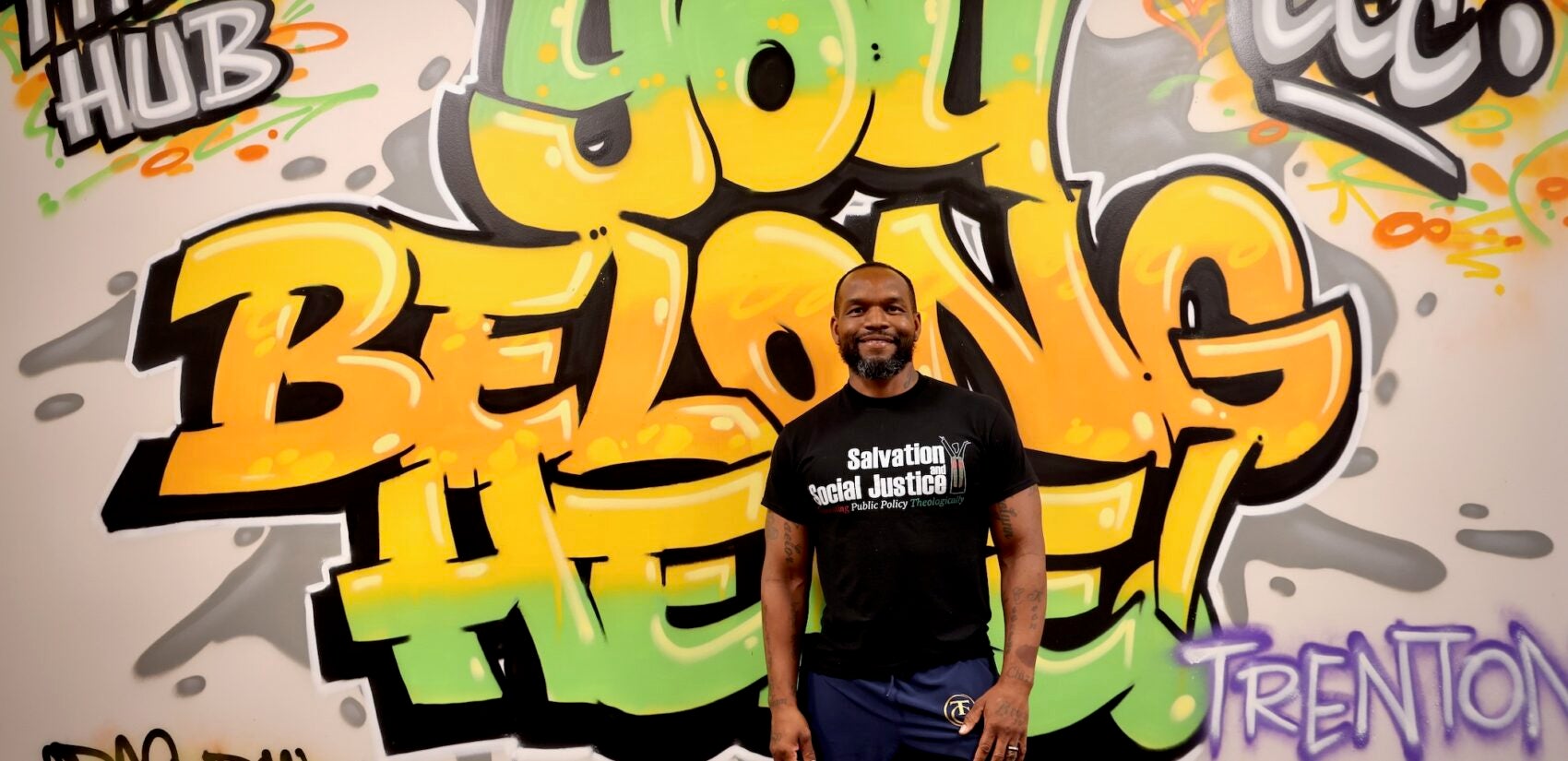 Dontae Thomas smiles in front of a mural reading YOU BELONG HERE!