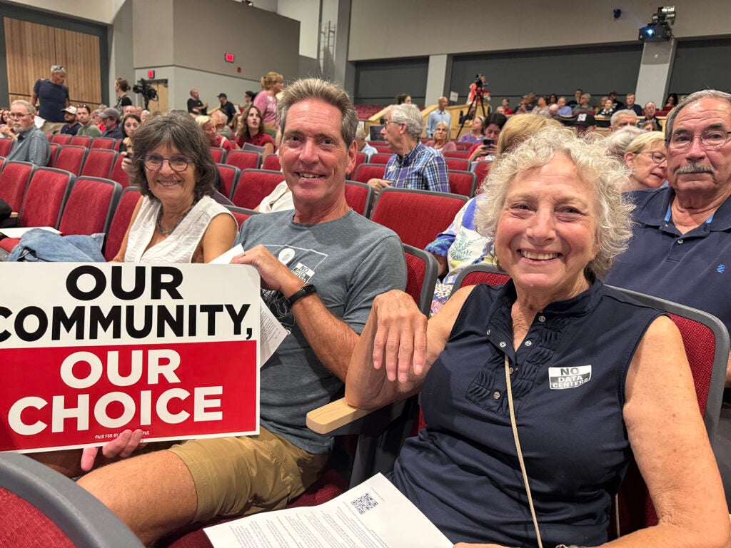 A man and a woman sit in an auditorium, with one holding a sign that reads "Our community, our choice"
