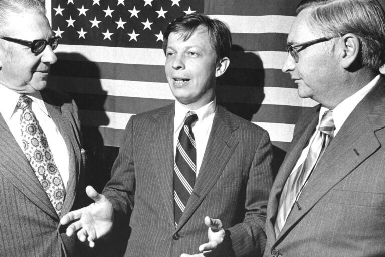 Franklin Kury speaking with 2 other men while standing in front of a large American flag.