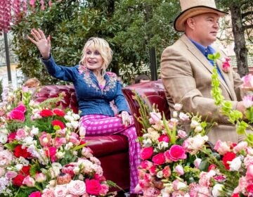 Dolly Parton waving to fans during a parade from a carriage, surrounded by pink and white flowers.