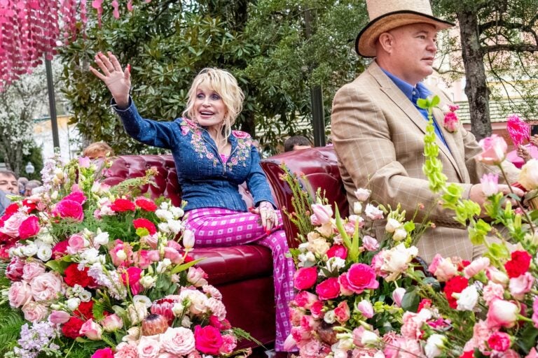 Dolly Parton waving to fans during a parade from a carriage, surrounded by pink and white flowers.