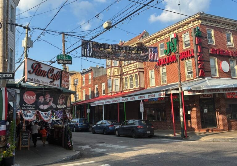 The beginning of the block of the Italian Market has restaurants lining both sides of the street, with lights and a banner hanging overhead saying 'Welcome to the Italian Market.'