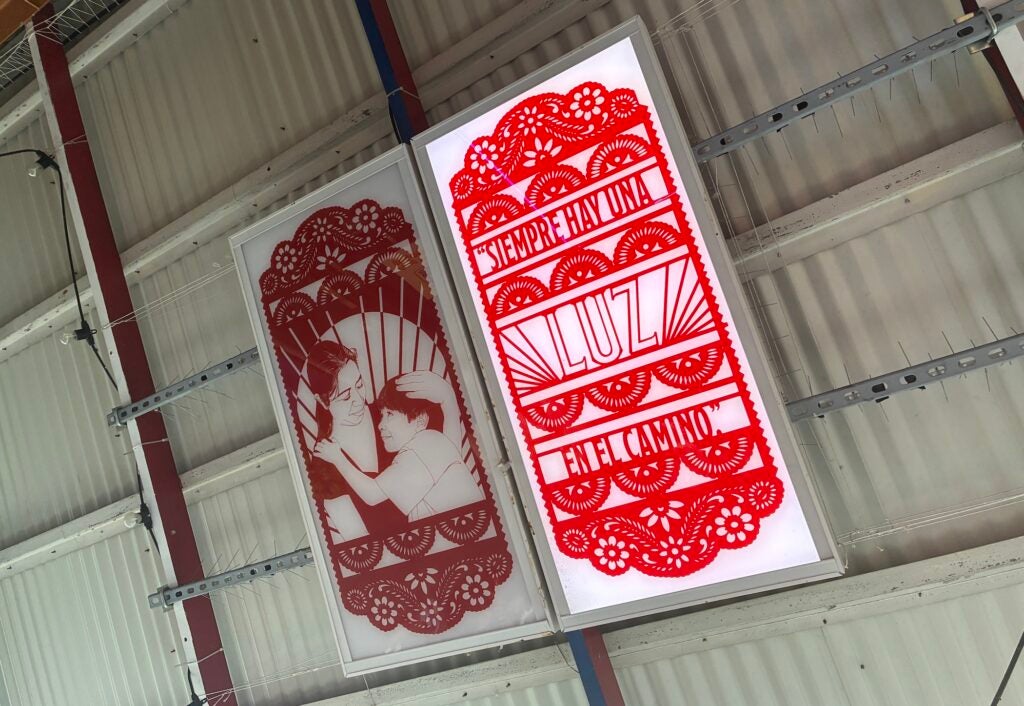 A light box is affixed to the ceiling above the entrance of a restaurant in the Italian Market. It is vibrant red, and says 'Siempre hay una luz en el camino."