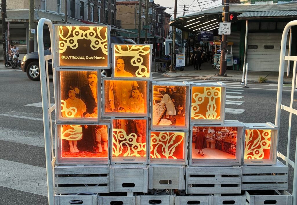 A movable push cart sits on the sidewalk in the Italian Market. It has wooden boxes many of which feature colorful images.