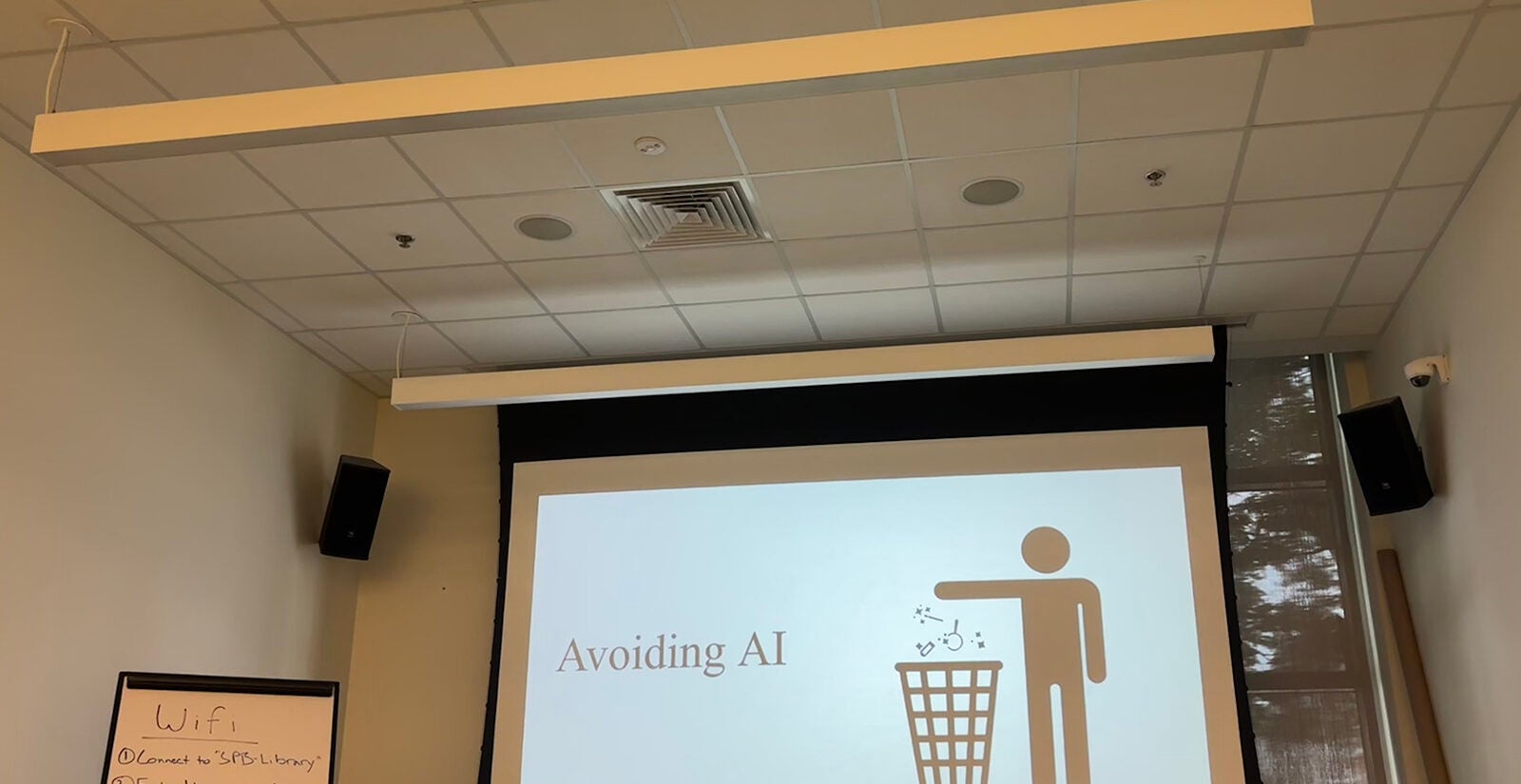 A projector shows a slide that reads "avoiding AI" and a person placing something in a trash can