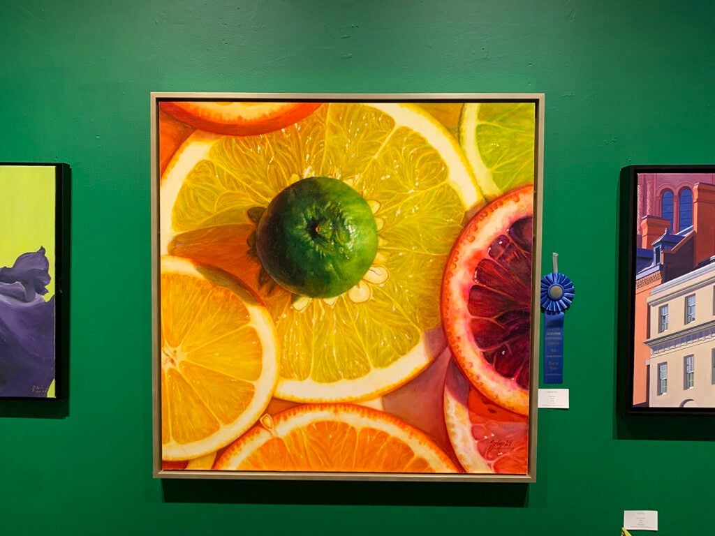 A large painting of an orange and other fruits hangs on a green wall between two other paintings