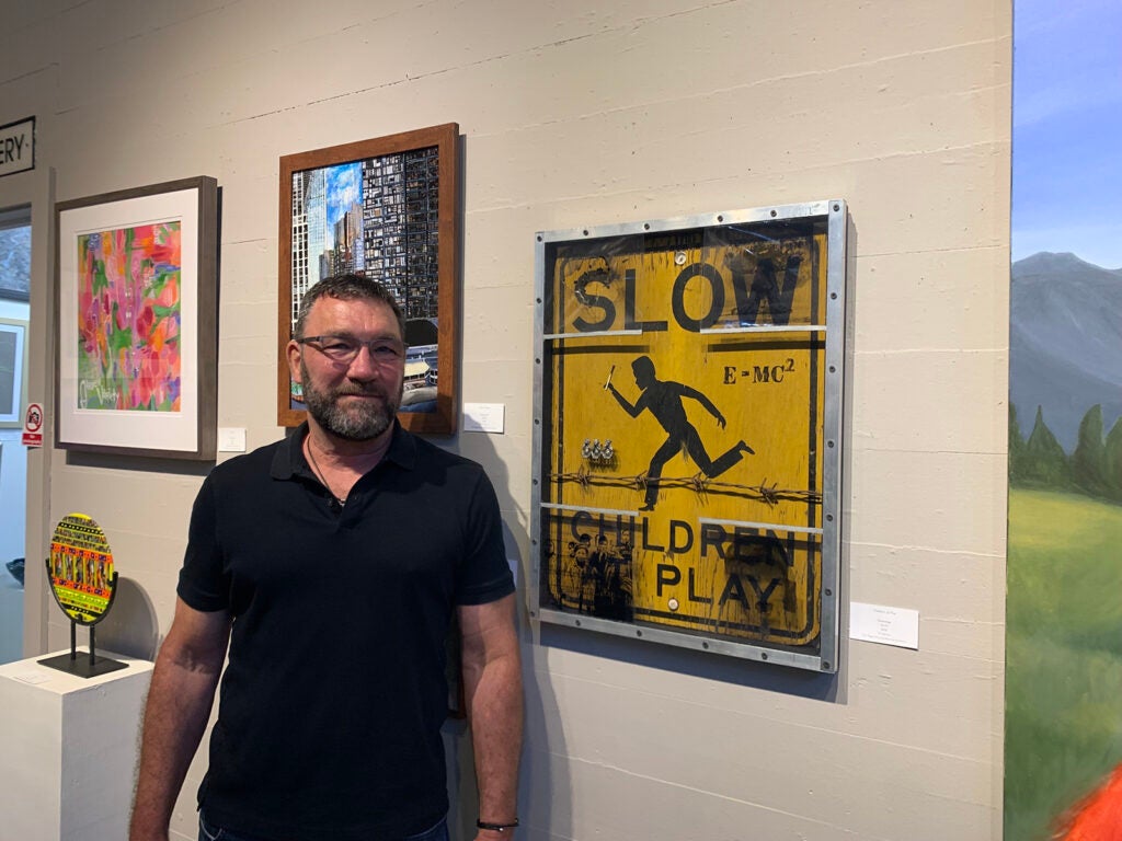 Tim Barton stands next to his art piece, which uses a yellow sign that reads "Slow/Children at play"