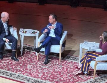 Cox-Shapiro-Garcia Josh Shapiro speaking into a microphone while sitting on stage at the Kimmel Center in white chairs with Spencer Cox and Lulu Garcia-Navarro.